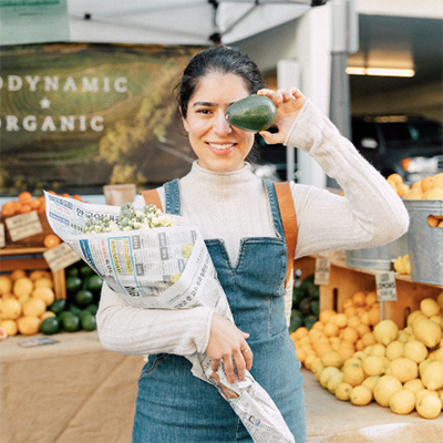 neeyaz with avocado held up to her eye/head for size and holding a bouquet of flowers at the market